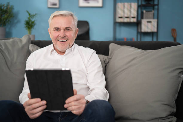 istock Smiling middle aged old man relaxing holding digital tablet reading e book sit on sofa at home, senior adult man enjoying using computer pad apps browsing internet shopping on couch in living room 1308644569