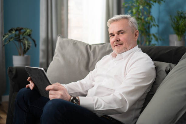 istock Smiling middle aged old man relaxing holding digital tablet reading e book sit on sofa at home, senior adult man enjoying using computer pad apps browsing internet shopping on couch in living room 1308644742