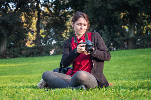 Girl sitting in the grass of the public park, photographing the surrounding landscape with her reflex camera. Casual, brunette.