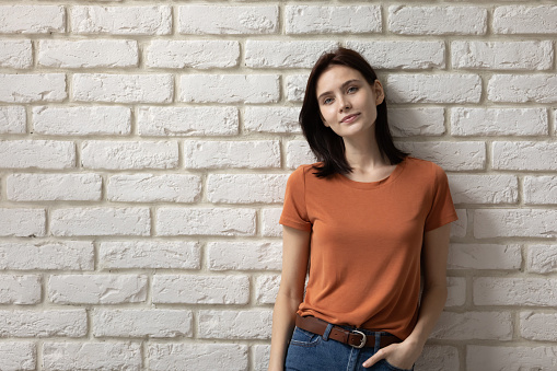 Portrait attractive young woman on white brick wall background, beautiful female wearing casual clothes orange t-shirt and blue jeans looking at camera, businesswoman or student posing for photo