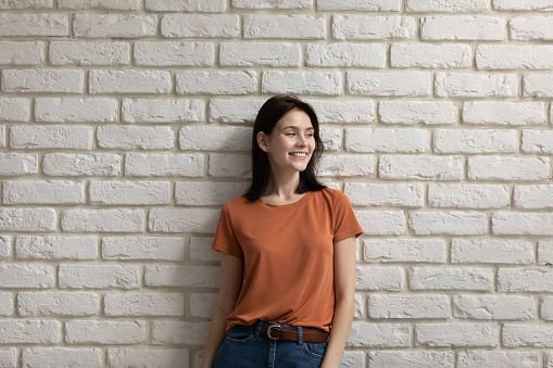 Portrait smiling attractive woman standing on white brick wall background, happy millennial female wearing casual clothes looking to aside, dreaming, visualizing good future, lost in thoughts