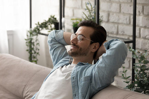 Close up calm man wearing glasses relaxing on cozy sofa, leaning back with hands behind head, taking nap, sleeping or daydreaming, enjoying lazy weekend, leisure time, breathing fresh air