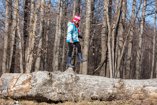 Side view of a young girl walking along a large fallen log in the woods. She just just about to step off a high point of the log and has one foot in mid-air.