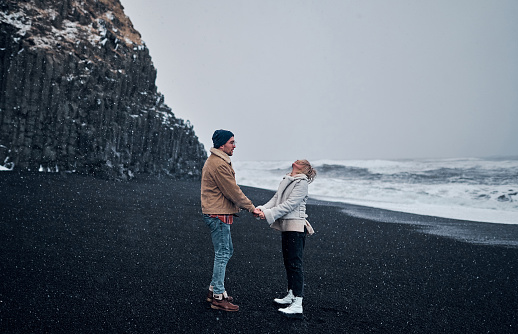 A loving couple of tourists holding hands look up at the falling snow, have fun walking along the beach with black volcanic sand and admire the great ocean waves. Travel, leisure, tourism.