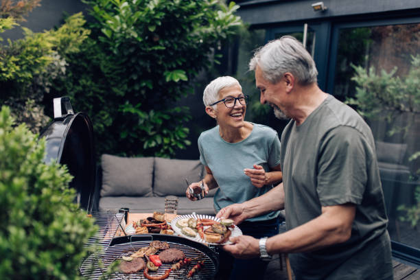 pareja mayor asando carne y disfrutando en el patio trasero - barbacoa fotografías e imágenes de stock