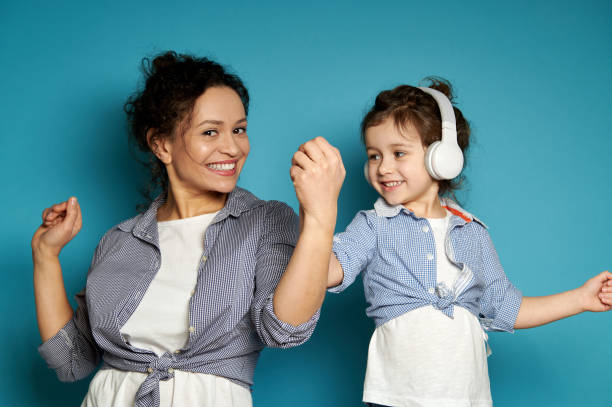 Beautiful mother and her daughter with headphones cutely smiling with toothy smile and looking at camera while posing over blue background. Beautiful mother and her daughter with headphones cutely smiling with toothy smile and looking at camera while posing over blue background with copy space cutely stock pictures, royalty-free photos & images