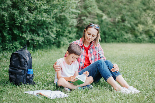 Young Caucasian mother reading book with son together. Family doing school homework in park outdoor. Mom helping child kid with education learning studying. Early development for children.