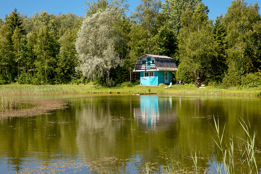 Cute blue color tiny house home by lake in summer. Beautiful rural scenic view and house reflection on lake.