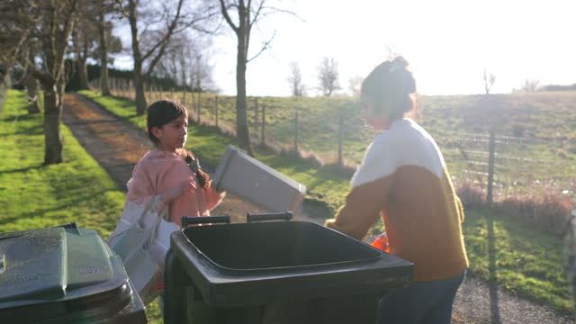 Aufnahme in voller Länge von zwei Schwestern, die Recyclinggegenstände ihre Einfahrt hinunter zu Mülleimern im Nordosten Englands tragen. Sie legten die Gegenstände in den Mülleimer, bevor sie zusammen weggingen.