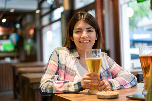 Young beautiful female customer enjoying a beer at the pub facing camera smiling - Lifestyles