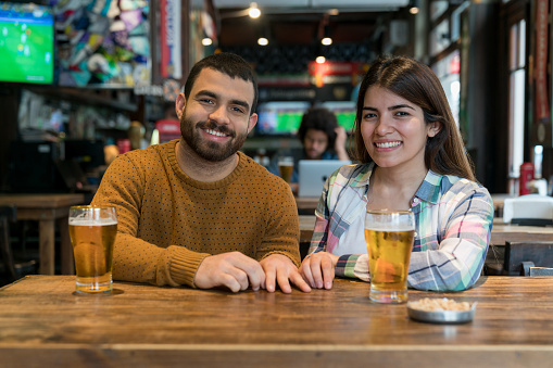 Happy young couple at the pub facing camera with a toothy smile while enjoying a beer - Lifestyles