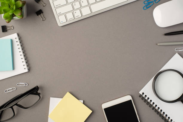 istock Overhead photo of notebook keyboard computer mouse magnifier pens phone plant glasses notes and paperclips isolated on the grey background with blank space 1308696992
