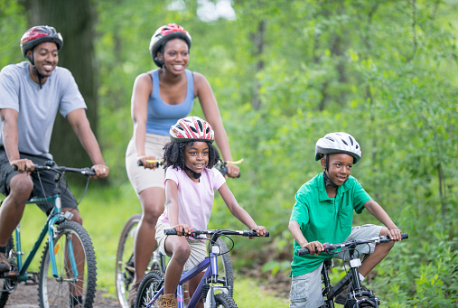 An active African American family with two children are biking outdoors together. The two children are riding their bikes in front of their parents. They are all happy to be spending time outdoors together.
