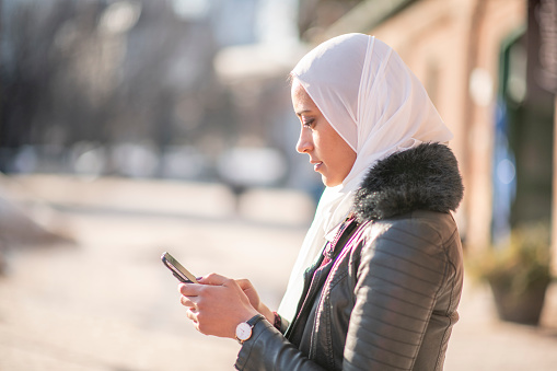 A side view of a Muslim woman wearing a hijab and is busy texting on her mobile phone.