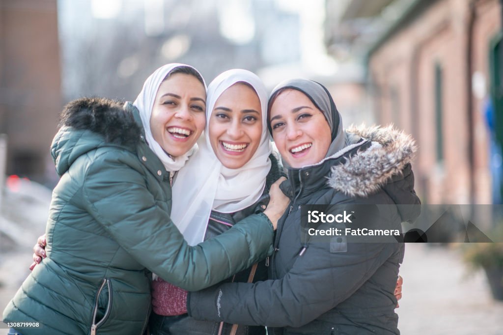 Three Beautiful Hijab Wearing Muslim Sisters Three beautiful Muslim sisters standing outdoors on a winter day. They are embracing and hugging each other for a photo. They are happy and smiling at the camera. Afghan Ethnicity Stock Photo