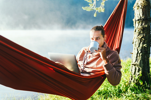 Young man in hammock working on his laptop and drinking hot coffee. Leisure activities / Remote working concept.