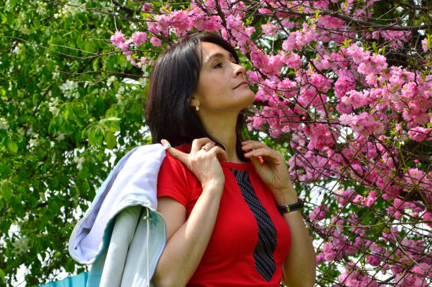 retrato de 45 y.o. mulher russa farejando galho sakura no fundo de macieira florescendo na primavera. - portrait adults apple tree cherry tree - fotografias e filmes do acervo