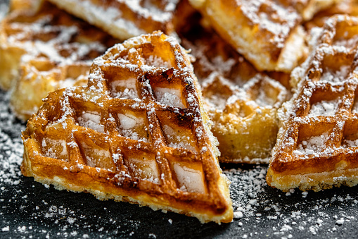 Close-up of fresh homemade waffle in heart shape. Dusted with icing sugar, and served on a black background. Format-filling macro with bright colors.