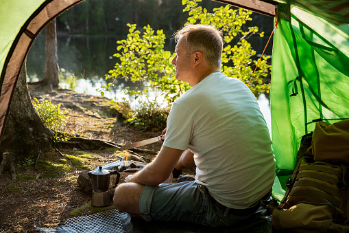 Man making coffee using espresso maker on campfire in forest on shore of a lake, sitting in tent, making a fire, grilling. Happy isolation concept. Exploring Finland. Scandinavian landscape.