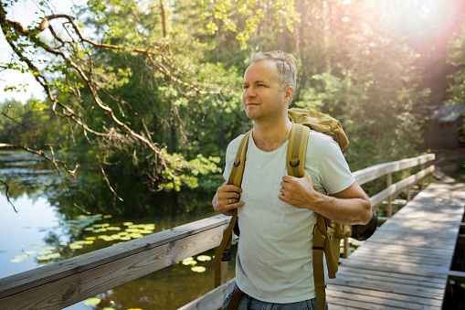 Mature man exploring Finnish nature in summer, walking across the bridge. Hiker with big backpack traveling in forests. Summer Scandinavian landscape of lakes and woods.