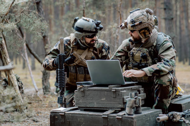 istock Bearded soldiers in uniform sit on military transport crates, analyze data on a laptop and work out tactics at a temporary forest base. In the background, you can see a soldier protecting the base 1308795536