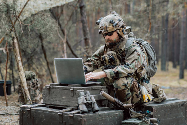 istock Bearded soldier in uniform sit on military transport crates, analyze data on a laptop and work out tactics at a temporary forest base. In the background, you can see a soldier protecting the base 1308795586