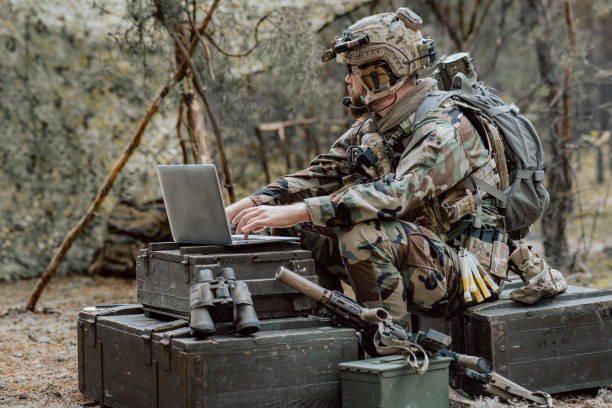 istock Bearded soldier in uniform sit on military transport crates, analyze data on a laptop and work out tactics at a temporary forest base. In the background, you can see a soldier protecting the base 1308795622