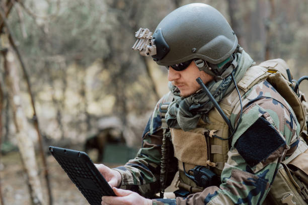 istock Bearded soldier in uniform sit on military transport crates, analyze data on a tablet and work out tactics at a temporary forest base. In the background, you can see a soldier protecting the base 1308796163