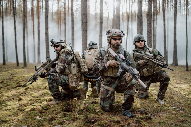 istock Four fully equipped, middle-aged soldiers in camouflage uniforms form a line, ready to fire, aiming with their rifles. A military operation in action, a unit standing in a dense forest 1308797591