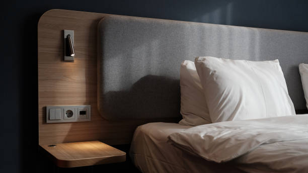 istock Close-up fragment of bedroom with empty bedside table, reading lamp and a USB socket in modern interior​ design home or hotel. Soft pillow and blanket, stylish comfortable furniture. Sun shadows. 1308815693