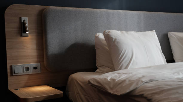 istock Close-up fragment of bedroom with empty bedside table, reading lamp and a USB socket in modern interior​ design home or hotel. Soft pillow and blanket, stylish comfortable furniture. Sun shadows. 1308815719