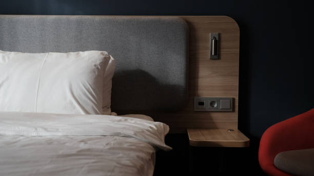 istock Close-up fragment of bedroom with empty bedside table, reading lamp and a USB socket in modern interior​ design home or hotel. Soft pillow and blanket, stylish comfortable furniture. Sun shadows. 1308815720