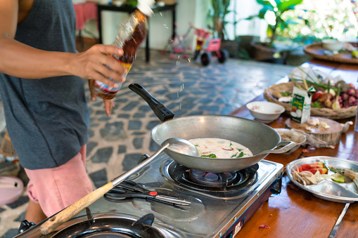 One Thai man is adding fish sauce to a traditional coconut milk soup called Tom Kha.  Cooking in a wok on a gas stove burner in his kitchen.  Three quarter length view of hime at the kitchen counter.
