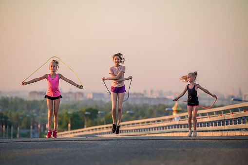Three slender sports girls jumping rope on the road on a summer evening against the backdrop of the city panorama and pink sunset sky