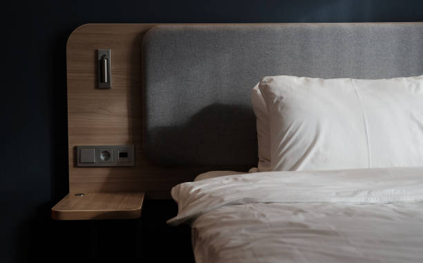 istock Close-up fragment of bedroom with empty bedside table, reading lamp and a USB socket in modern interior​ design home or hotel. Soft pillow and blanket, stylish comfortable furniture. Sun shadows. 1308819014
