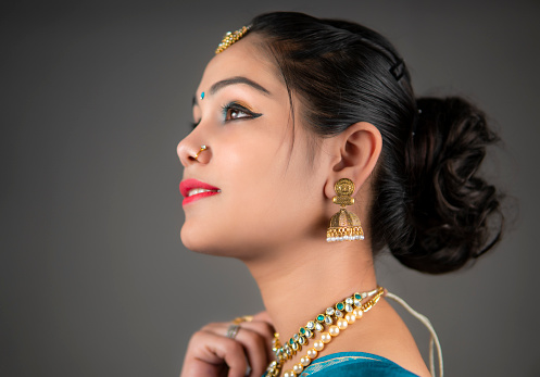 Side view close-up portrait of beautiful, happy Indian woman in traditional sari and gold jewelry looking away with a smile and thinking. She is standing against a gray background in studio light.