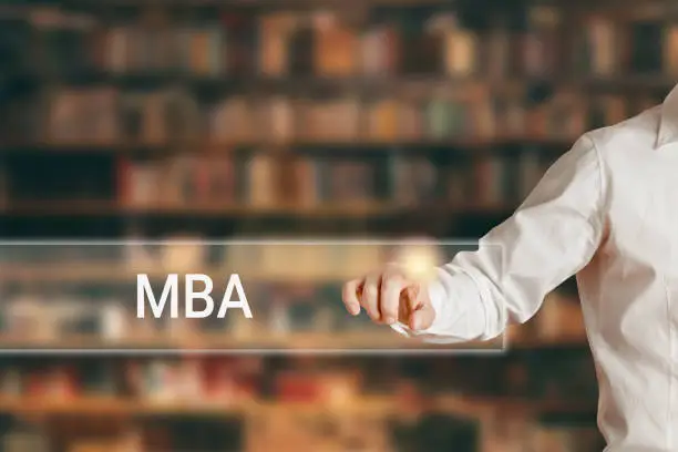 Photo of Male hand pressing the word MBA on a virtual search display bar against library background.