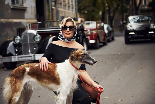 Blonde woman in sunglasses and in black dress near old vintage classic car with her dog.