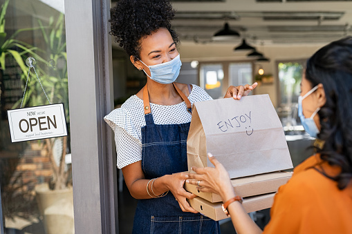 African restaurant owner working only with take away orders during corona virus outbreak. Young black woman wearing face mask giving takeout meal to customer outside her cafeteria. Customer pick up take-away food ordered at home, support local small business during covid19 pandemic.