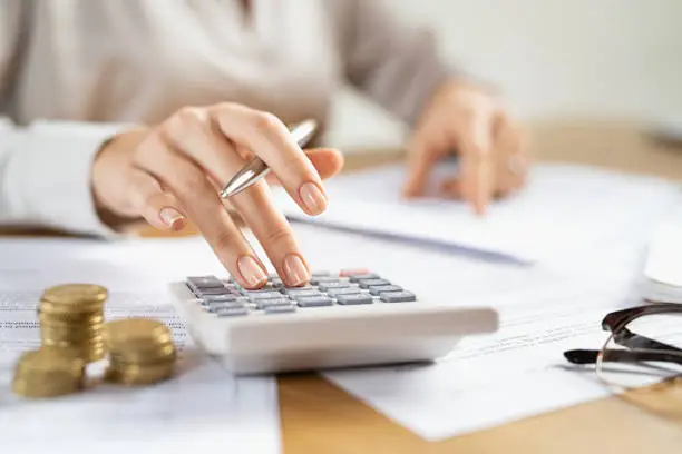 Woman using a calculator doing a balance of monthly expenses. Close up of businesswoman working with financial data and using calculator with coins on desk at office. Deatil of hands calculating home finances and annual taxes.