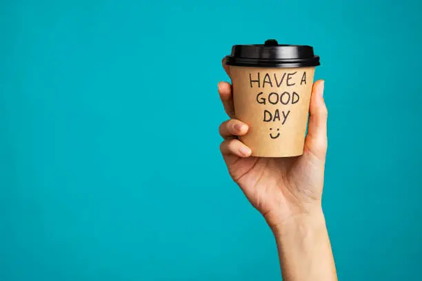 Close up of hand of young woman holding take away coffee cup with text written on it. Woman hand holding a coffee paper cup isolated on blue background. Morning disposable mug with message of good day written over it.