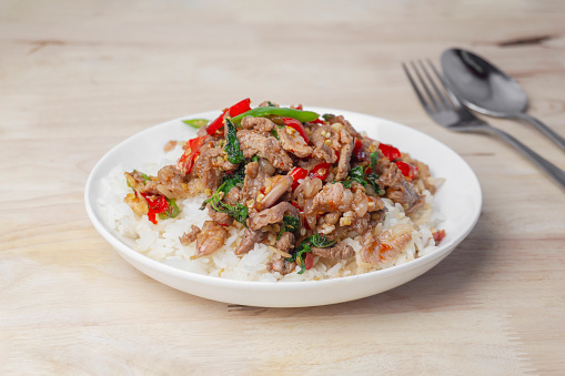 Close up Rice topped with stir-fried beef and holy basil (Pad Ka Prao) on the wooden table with stainless spoon and fork popular Thai street food.