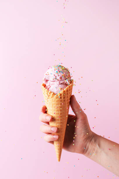 Female hand holding the pink ice cream with falling sprinkles in waffle cone Female hand holding the pink ice cream with falling sprinkles in waffle cone on pink background ice cream cone photos stock pictures, royalty-free photos & images