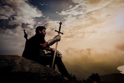 Medieval fantasy knight sitting relaxed during the sunset in the mountains looking thoughtful into the distance. Creative dramatic color retouching with added grain.Part of an E+ and S+ series.