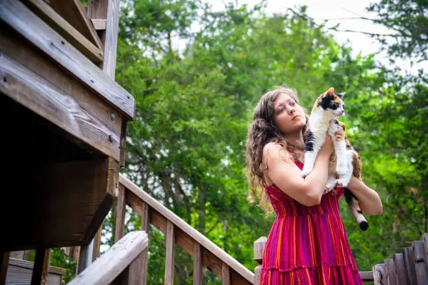 Happy smiling young woman bonding holding in hands calico cat pet companion, friends showing affection outside outdoors in home garden on wooden deck stairs