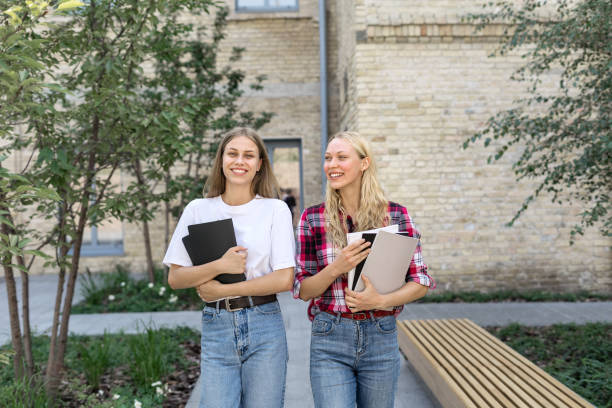 Two happy and smiling young academy student girl walking outdoors after study, holding copybooks in hands, spending time near campus together, smiling wide, talking to each other