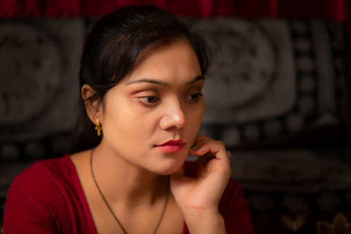Indoor low key portrait of beautiful, sad, serene married Indian woman sitting alone at home near the sofa and thinking deeply with a blank expression.