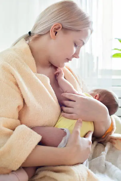 Baby eating breast milk. Caucasian blonde young mother breastfeeding her baby at home vertical photo. Concept of lactation.