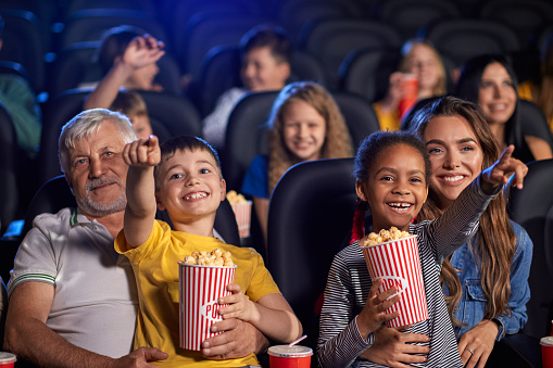 Front view of multiracial kids holding popcorn and sitting on knees of parents. Little african girl with caucasian woman and caucasian boy with senior grandfather. Family, entertainment concept.