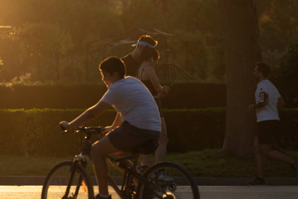 Madrid, Spain - June 20, 2020: A boy passes quickly, riding a bicycle, in front of a couple who chat and practice skating through the Rosaleda area, in the Retiro Park.